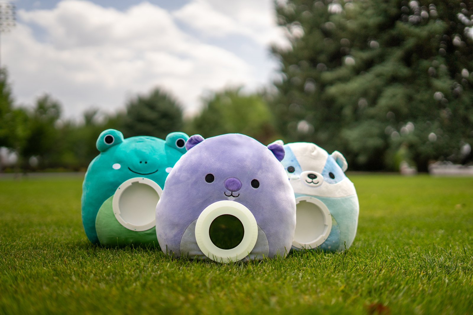 Three Merfi plush companions — a frog, a bear, and a husky — sitting on the grass, each with a round screen built into its belly