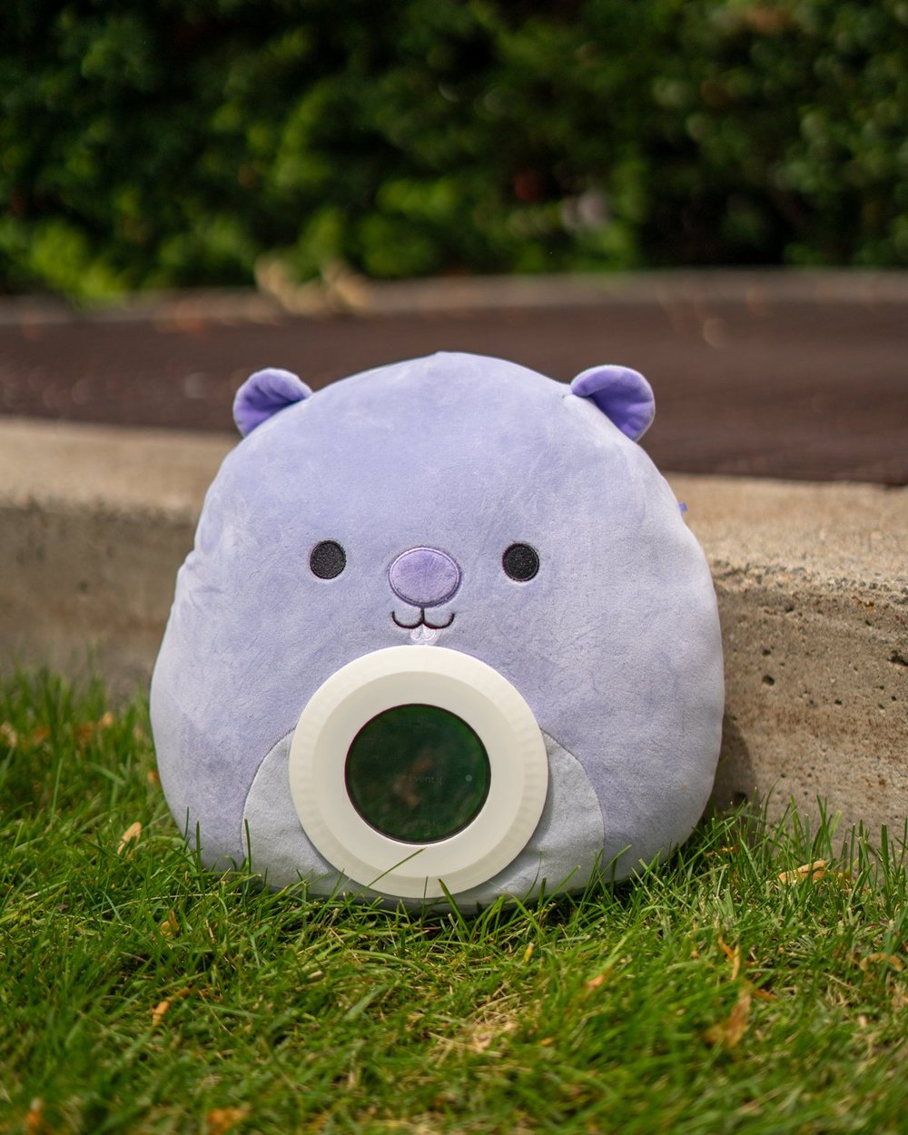 A soft purple Merfi bear sitting on the grass with its rounded screen in front